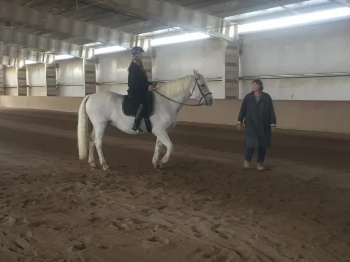 Instructor coaching a rider on a white horse from the ground during an indoor arena lesson at Mountain View Ranch